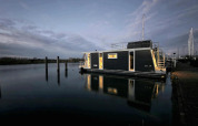 Casa flotante Havenlodge Lelystad en Marina Parcs Lelystad, Países Bajos, iluminada al anochecer sobre agua tranquila.