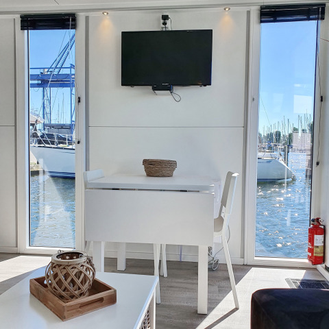 Interiør af houseboat Havenlodge Lelystad med bord, stol, tv og udsigt over marinaen i Nederlandene.