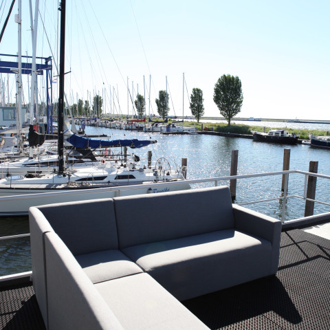 Blick von einem Hausboot bei Havenlodge Lelystad auf eine graue Couch, Segelboote und Yachthafen.