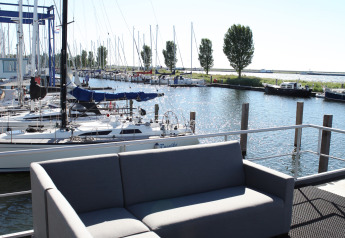 View from a houseboat at Havenlodge Lelystad, with a grey sofa, marina and sailboats in sunlight.