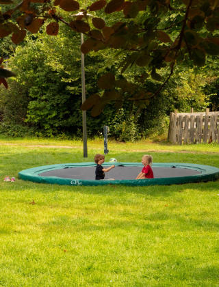 Dos niños juegan en una cama elástica enterrada, rodeados de césped, en Feather Down Het Boshuis, Gelderland.