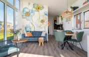 Interior moderno y luminoso de una cabaña con sofá azul, mural floral y zona de comedor junto al agua en Knilles.