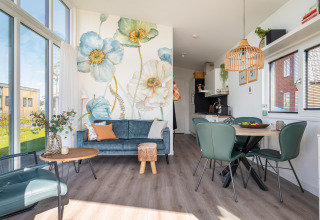 Intérieur lumineux d’un lodge moderne avec mur fleuri, canapé bleu et coin repas au bord de l’eau à Knilles.