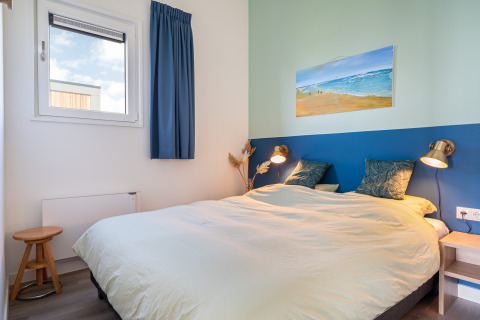 Dormitorio luminoso con cama doble, cortinas azules, lámparas modernas y cuadro de playa en la pared.