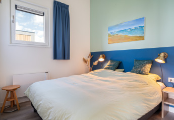 Dormitorio luminoso con cama doble, cortinas azules, lámparas modernas y cuadro de playa en la pared.