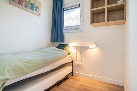 Lichte, kleine slaapkamer met eenpersoonsbed, raam, blauw gordijn en wandkast in de Knilles Lodge.