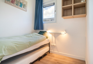 Lyst lille værelse med enkeltseng, vindue med blå gardiner, væghylde og blødt lys på Knilles-lodgen.