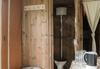 Baño rústico con paredes de madera en una cabaña, con inodoro, cubo de basura, tetera y vasos.