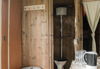 Baño rústico con paredes de madera en una cabaña, con inodoro, cubo de basura, tetera y vasos.