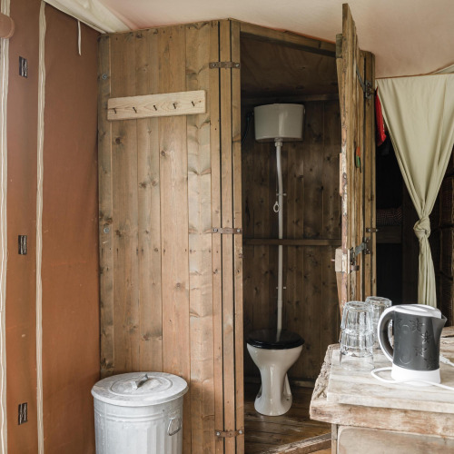 Baño rústico con paredes de madera en una cabaña, con inodoro, cubo de basura, tetera y vasos.