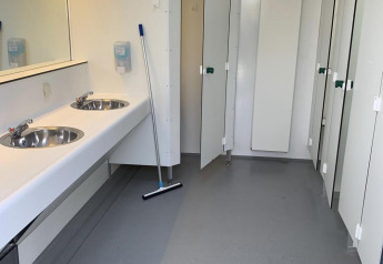 Baño público limpio con lavabos, espejo, herramienta de limpieza y cabinas cerradas en un parque holandés.