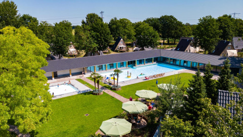 Vista aerea di un villaggio tiny house con piscina centrale, ampio prato verde e case immerse tra gli alberi.