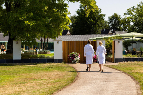 Dos personas con albornoces caminan hacia una tiny house rodeada de jardín, árboles y zona de bienestar.
