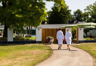 Dos personas con albornoces caminan hacia una tiny house rodeada de jardín, árboles y zona de bienestar.