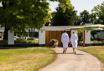 Dos personas con albornoces caminan hacia una tiny house rodeada de jardín, árboles y zona de bienestar.