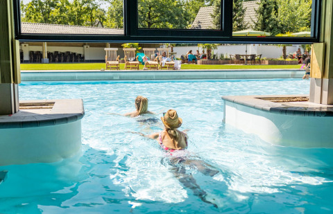 Dos personas nadan de una piscina interior a una exterior en una tiny house, con gente relajándose afuera.