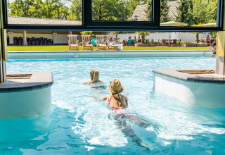 Dos personas nadan de una piscina interior a una exterior en una tiny house, con gente relajándose afuera.