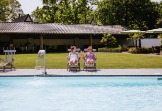Dos personas relajándose en tumbonas junto a una piscina frente a una tiny house en un día soleado.