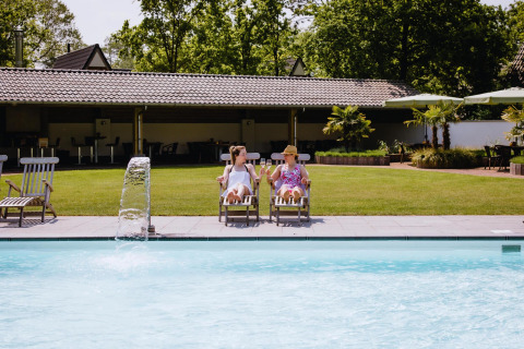 Two people relaxing on lounge chairs by a swimming pool in front of a tiny house on a sunny day.