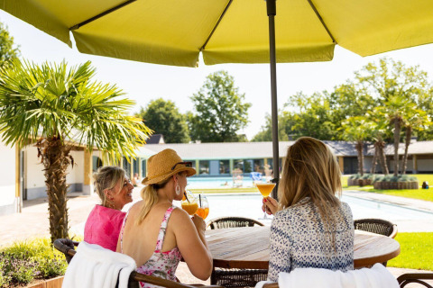 Three women enjoy cocktails under a large umbrella by a tiny house pool, tropical trees in the background.