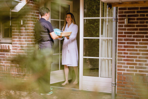 A woman in a bathrobe receives a basket from a person at the door of a tiny house with brick walls.