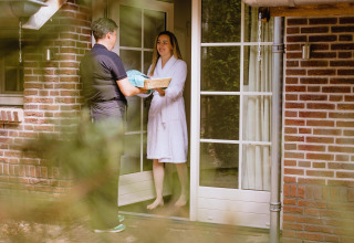 A woman in a bathrobe receives a basket from a person at the door of a tiny house with brick walls.