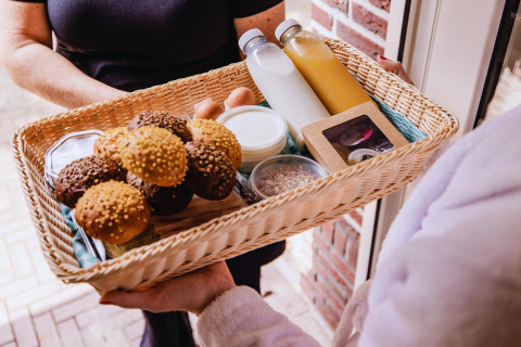Cestino colazione con panini, latte, succo, uova e yogurt consegnato alla porta di una tiny house.