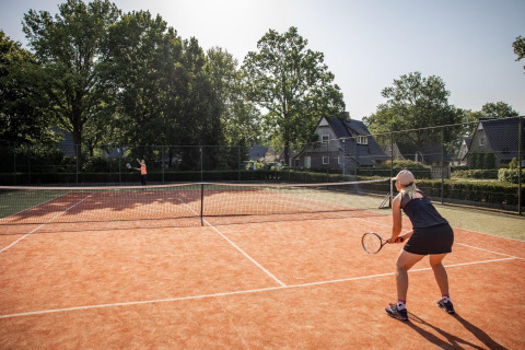 Dos personas juegan al tenis en una pista al aire libre con casas pequeñas y árboles de fondo en un día soleado.