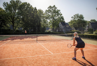 Two people play tennis on an outdoor court with small houses and trees in the background on a sunny day.