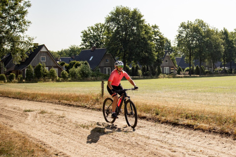 Person cykler på en grusvej foran små huse og græsareal på en solrig dag i landet.