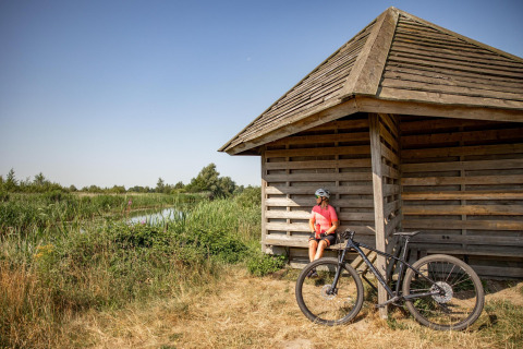 Ciclista con casco descansa junto a una pequeña casa de madera cerca de un estanque y vegetación.