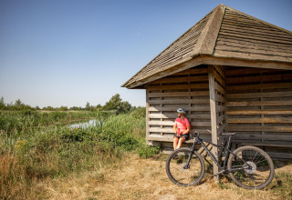 Ciclista con casco descansa junto a una pequeña casa de madera cerca de un estanque y vegetación.