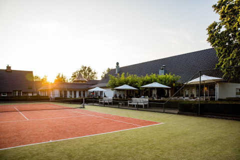 Pista de tenis al aire libre frente a una tiny house, con mesas, sombrillas y el atardecer de fondo.