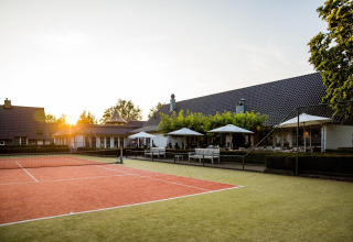 Pista de tenis al aire libre frente a una tiny house, con mesas, sombrillas y el atardecer de fondo.