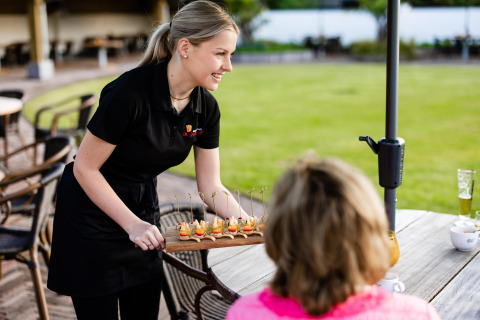 A waitress serves appetizers to a guest at an outdoor table near a tiny house and a green lawn.