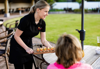 Une serveuse sert des amuse-bouches à une cliente à une table extérieure près d’une tiny house verdoyante.