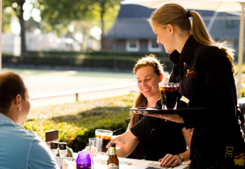 Een serveerster serveert drankjes aan twee lachende gasten op het terras van een tiny house in de zon.