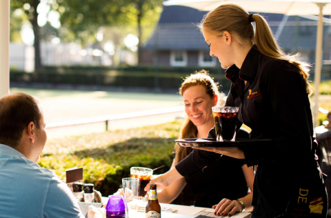 A waitress serves drinks to two smiling guests seated outdoors at a small house on a sunny day.
