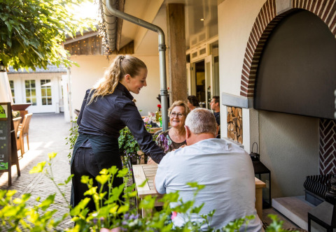 Una camarera sirve comida a una pareja sonriente en la terraza de un pequeño restaurante casero.