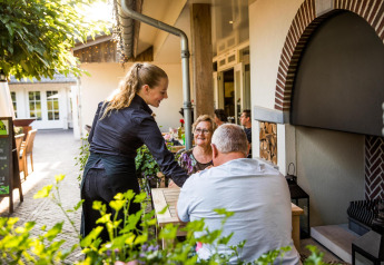 Une serveuse apporte des plats à un couple assis en terrasse devant une petite maison accueillante.