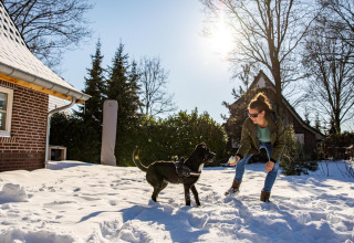 Frau spielt mit schwarzem Hund im Schnee vor dem Wellness Lodge XL am Hof van Salland, Niederlande, an einem sonnigen Tag.