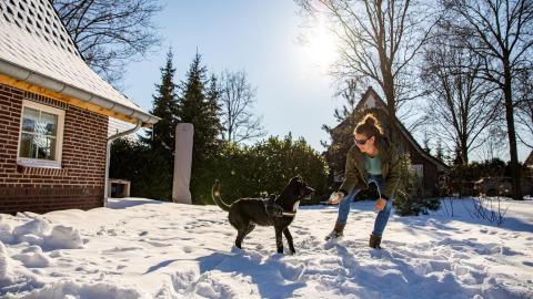 Mujer juega con un perro negro en la nieve, frente a Wellness Lodge XL en Hof van Salland, Países Bajos, en un día soleado.