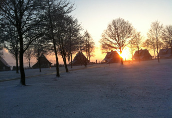 Amanecer sobre casas pequeñas en un paisaje invernal nevado, rodeadas de árboles sin hojas.