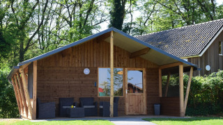 Baita in legno al Camping de Koeksebelt nei Paesi Bassi, terrazza e sedie, circondata da alberi.