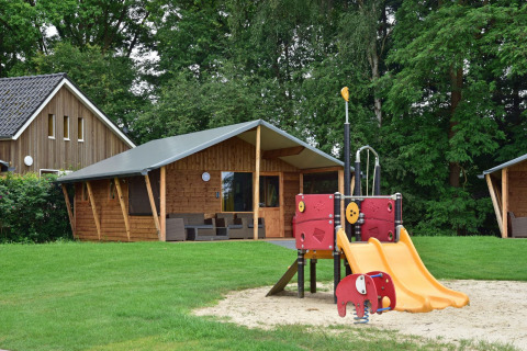 Parco giochi con scivolo davanti a baite in legno al Cabin di Camping de Koeksebelt nei Paesi Bassi, vista bosco.