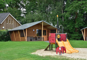 Parco giochi con scivolo davanti a baite in legno al Cabin di Camping de Koeksebelt nei Paesi Bassi, vista bosco.