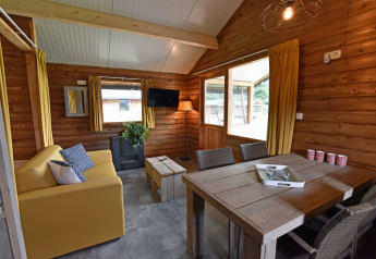 Indvendig udsigt over en hyggelig træhytte med gule gardiner, sofa, spisebord og store vinduer.