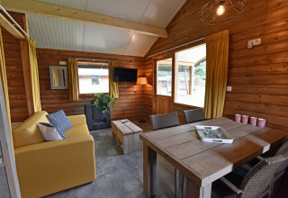 Indvendig udsigt over en hyggelig træhytte med gule gardiner, sofa, spisebord og store vinduer.