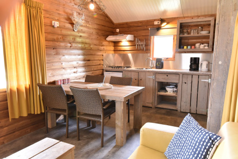 Interiør af en hyggelig træhytte med spisebord, køkken og store gule gardiner på Camping de Koeksebelt.