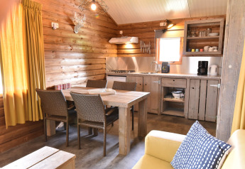 Gezellige houten blokhut met eettafel, keuken en gele gordijnen op Camping de Koeksebelt in Nederland.